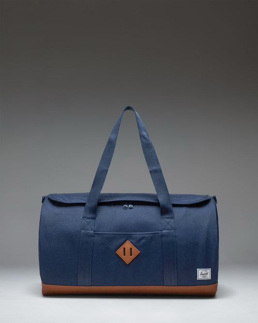 Herschel Heritage Duffle bag in Navy Blue with Saddle Brown handles and detailing, shown from a three-quarter angle on a white background.