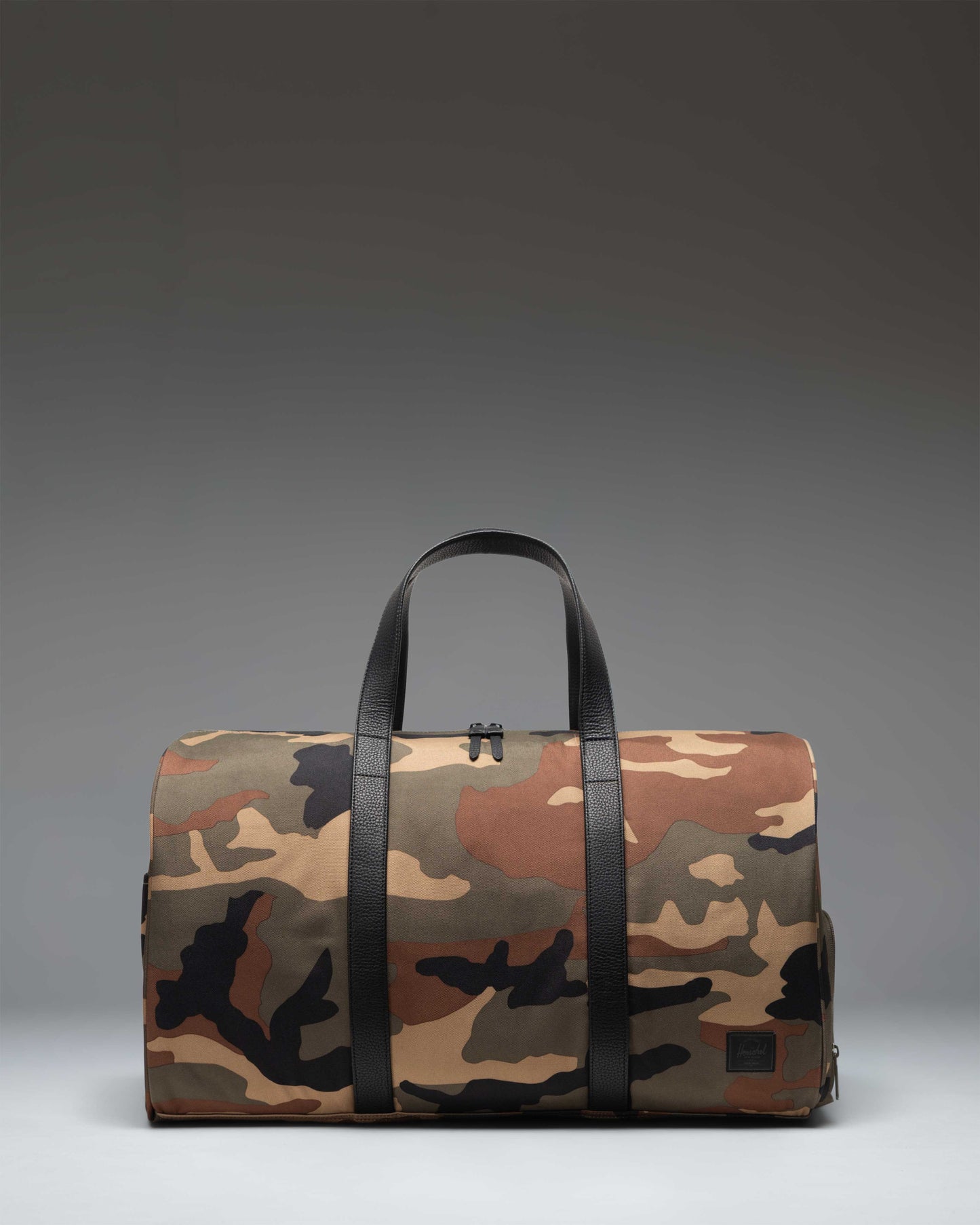 Herschel Novel Duffle bag in Woodland Camo with black handles and a Black Label logo on the front.