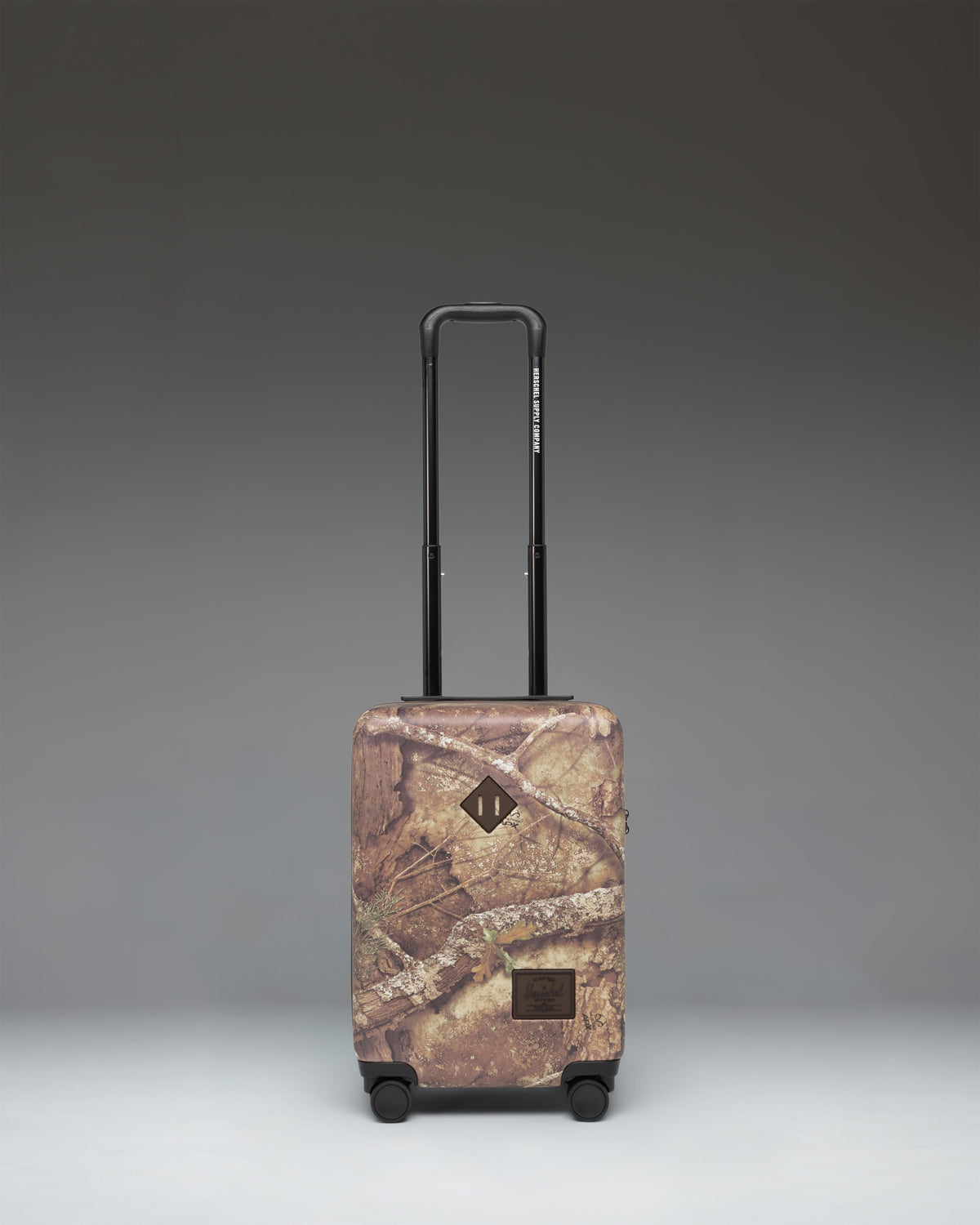 Herschel Heritage Carry On Luggage in Realtree APX Camo pattern, showing the front view with a black zipper and signature logo label.