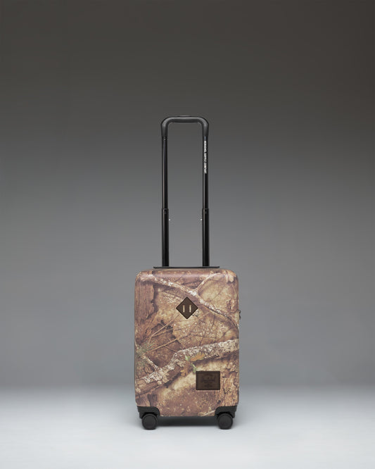 Herschel Heritage Carry On Luggage in Realtree APX Camo pattern, showing the front view with a black zipper and signature logo label.